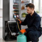 Técnico inspecionando a parte traseira de uma geladeira com cilindro de gás e manômetro em uma cozinha moderna.
