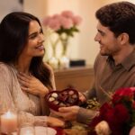 Um casal sentado junto em uma mesa aconchegante, trocando gestos carinhosos em um ambiente romântico com velas e flores.